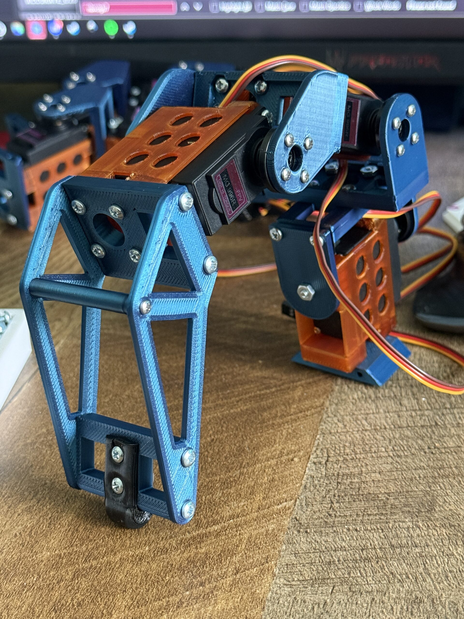 A high-angle, close-up photograph shows a fully assembled, 3D-printed hexapod leg standing on a wooden surface. The leg is composed of three main sections: a base joint, a middle "thigh" segment, and a long, tapered lower leg (tibia).