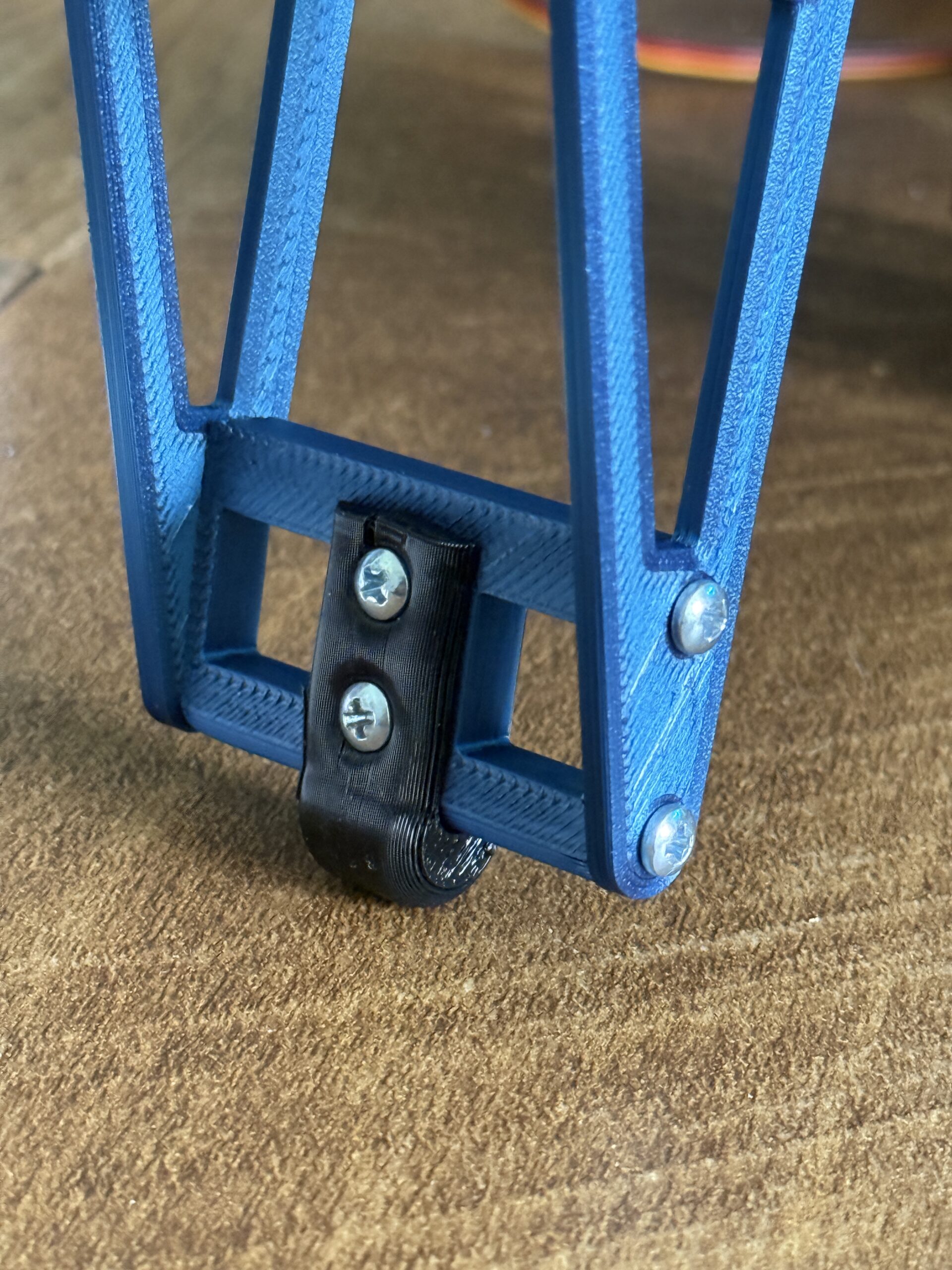 A macro photograph focusing on the tip of a 3D-printed hexapod leg. The lower segment is a light-blue, open-frame tibia with a triangular truss design, held together by silver Phillips-head screws. At the very tip, a small, curved black 3D-printed foot pad is securely attached with two vertical screws.